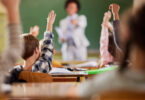 Students raising their hands in a classroom as a teacher stands at the front near a green chalkboard. (Getty Images)