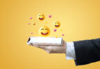 A hand holding a cell phone with emojis popping out of the screen.